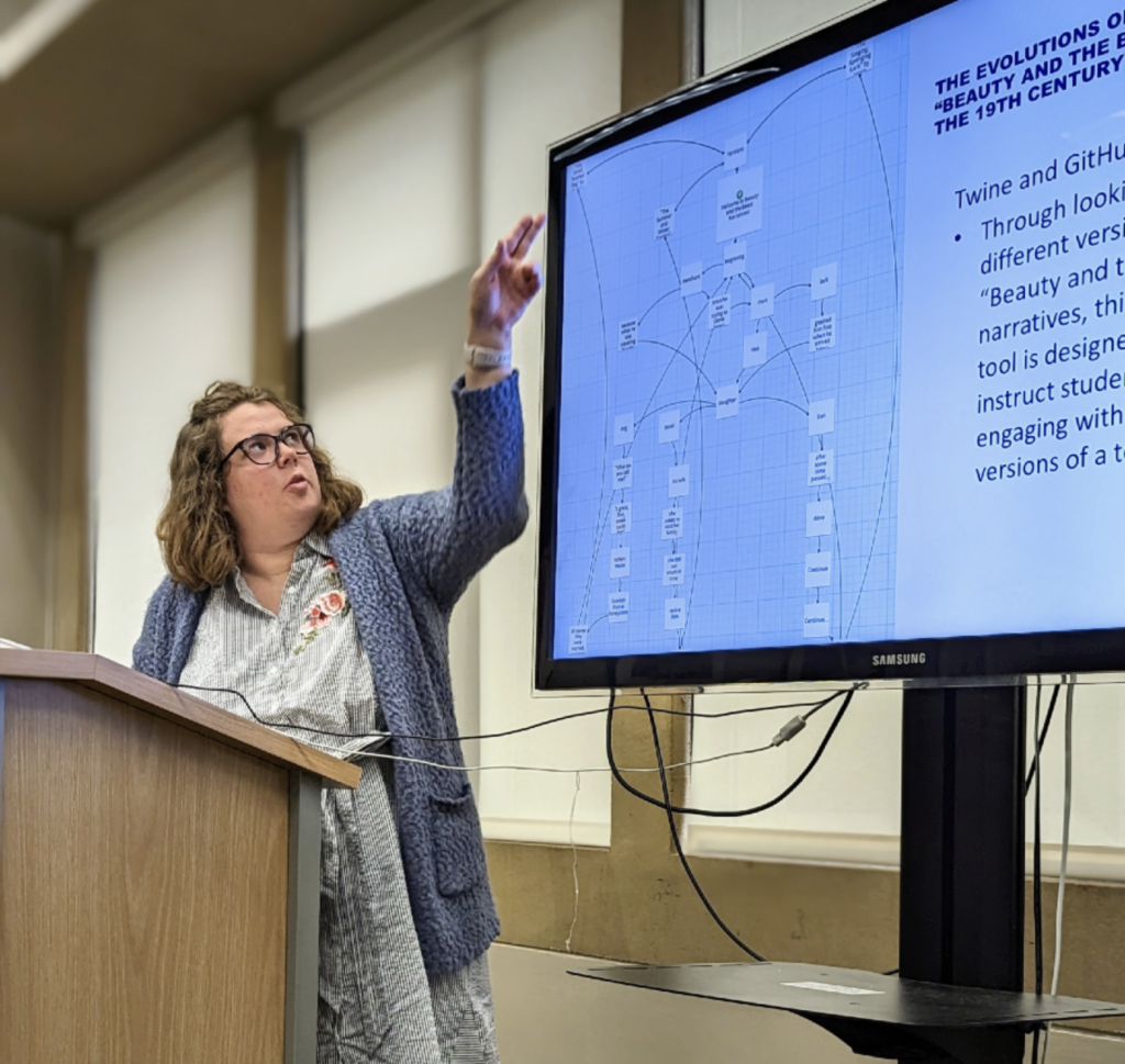 A woman in glasses stands at a podium and points to a presentation on a screen. The display shows a digital flowchart graphic on the left and text about her DH project on the right.