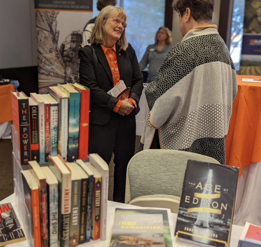 In the background, two women talk to one another (one has her back to the camera). In front of them is a table with a book display of books related to the field of energy humanities.