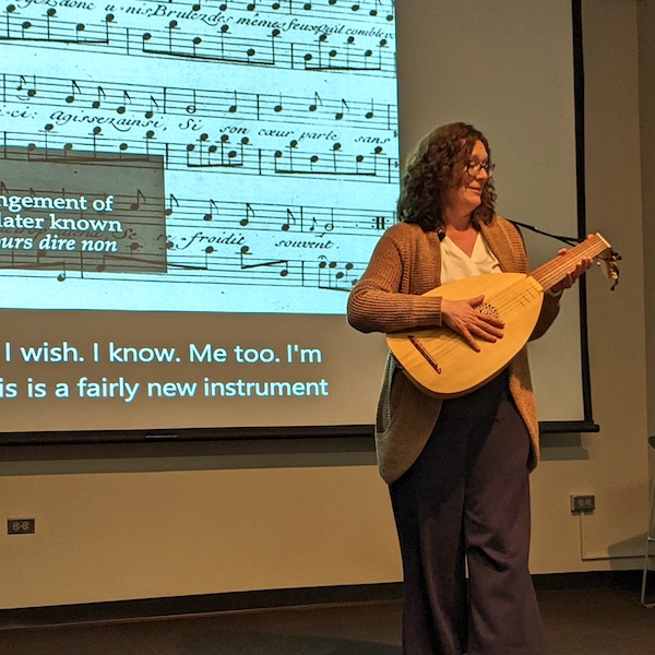 A white woman plays the lute in front of a screen displaying early modern musical notation