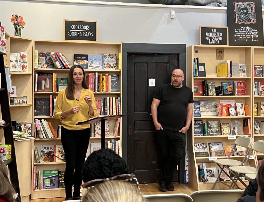 A white woman with long, straight brown hair stands at a podium in a bookstore. She is wearing black pants and a yellow blouse. To one side, a bald, bearded white man in black leans against a bookshelf listening to her speak. The backs of the audience's heads are just visible.