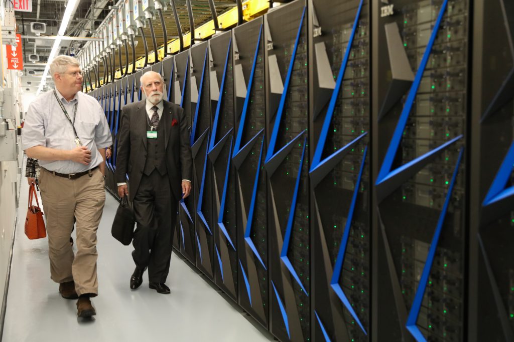 Two men - one dressed in khaki pants and a light-colored shirt and the older one dressed in a black suit, walk down a hallway filled by an ORNL supercomputer