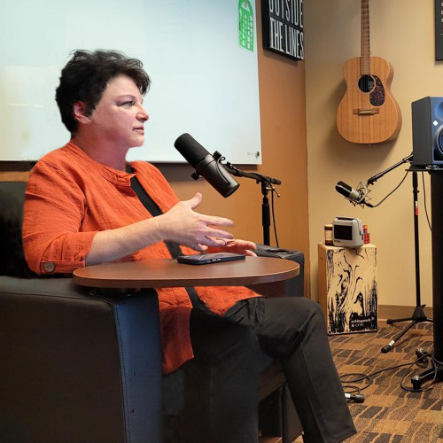 A middle-aged woman with short brown hair and wearing a dark orange dress jacket sits in a chair in front of a microphone. On the wall behind her, a guitar hands on the wall of a studio.