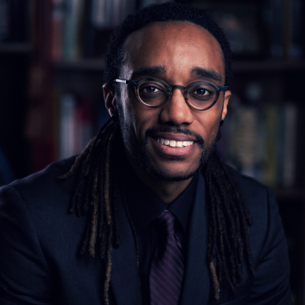 A Black man with glasses and long dreadlocks smiles at the camera. He is wearing a tie and dark blue suit jacket.