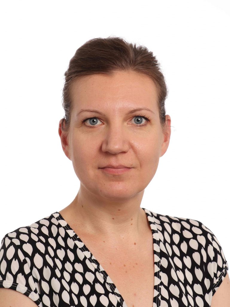 A headshot photo of a white woman in a v-necked black shirt with a cream-colored spot pattern on it. Her brown hair is pulled back on her head.