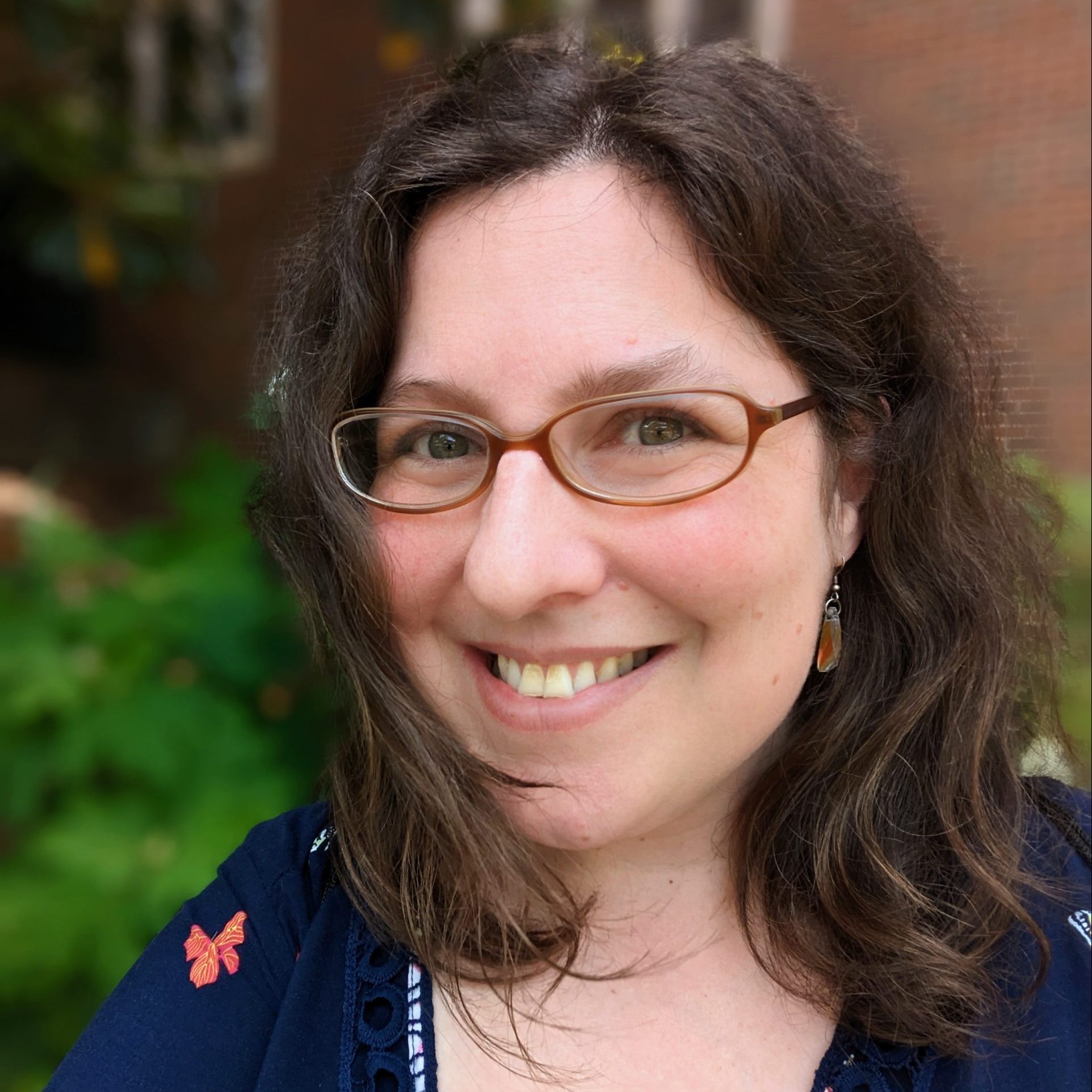 A white woman with windswept brown hair and glasses smiles at the camera outside of a brick building with green shrubbery.