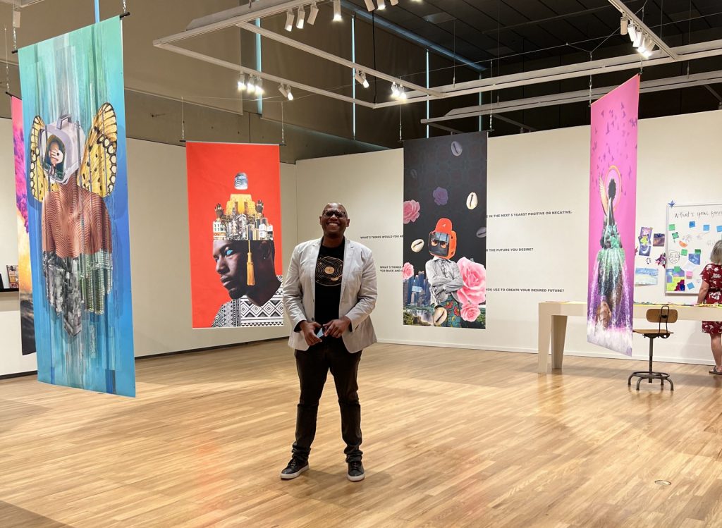 A Black man dressed in black with a tan blazer stands in the middle of a large room with pale wood floors. He is surrounded by four hanging banners with Afrofuturist art on them.