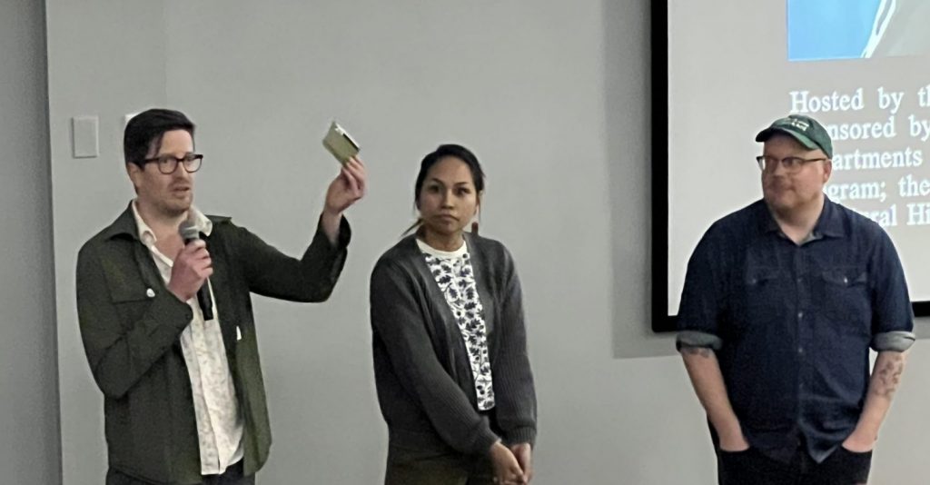 Cropped photo of two men and one woman standing in front of a projection screen. The man on the left is holding up something small in his hand and speaking into a microphone while the other two look at him.