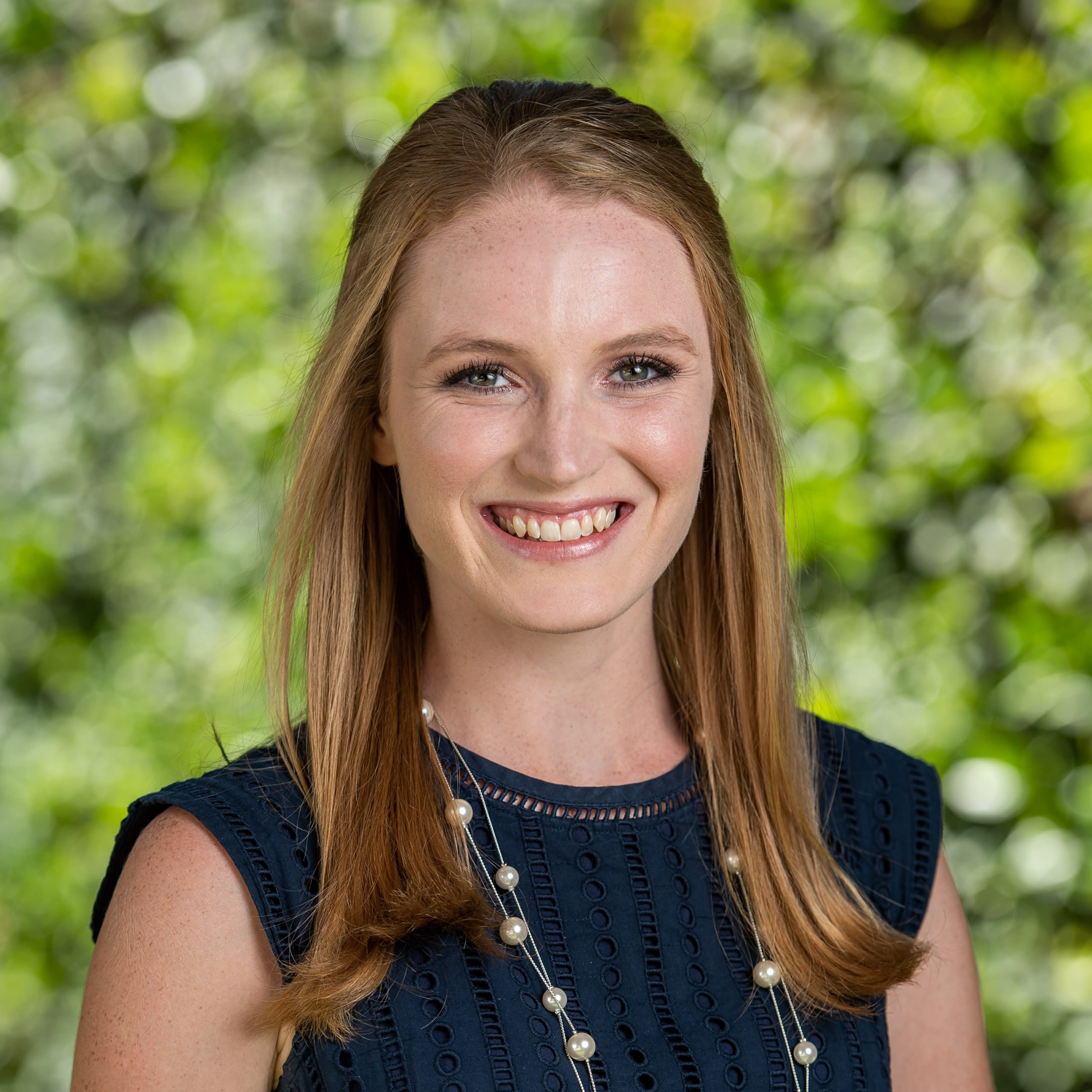 A white woman in a sleeveless black dress with a necklace and long blonde hair smiles at the camera. She is outside in front of a backdrop of greenery.