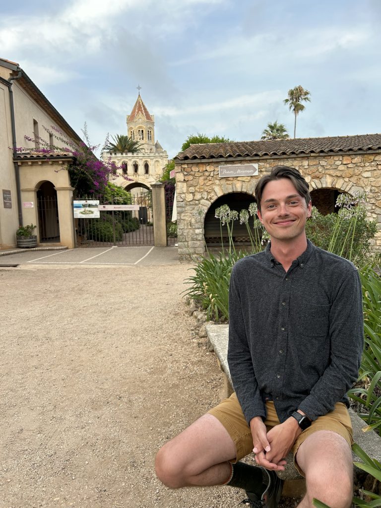 A young man sits on a stone bench smiling at the camera. Behind him is raised-bed garden and a courtyard leading to several old-fashioned stone buildings.