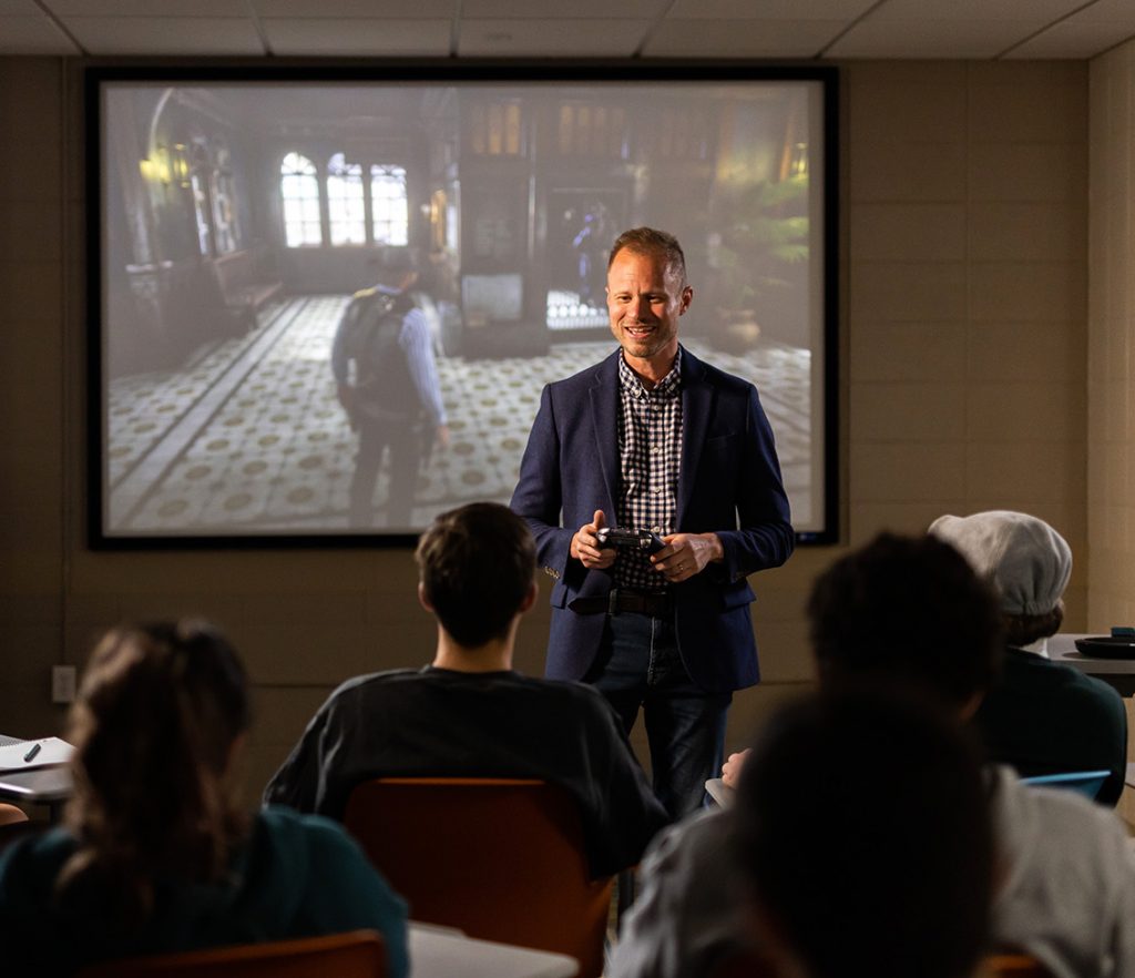 A white man in a blue suit stands at the front of a classroom speaking to a room full of students. Behind him on a screen is a still image from a video game.