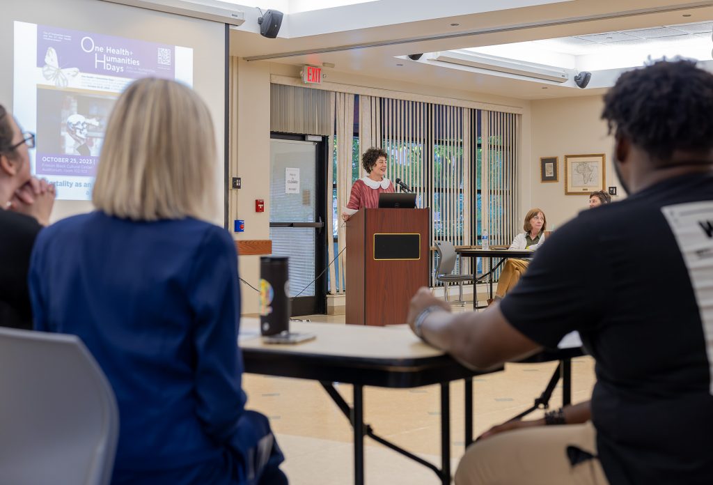 A white woman with short curly dark hair stands behind a lectern in front of a projection screen. Seated at tables around her is an audience viewed from the back.