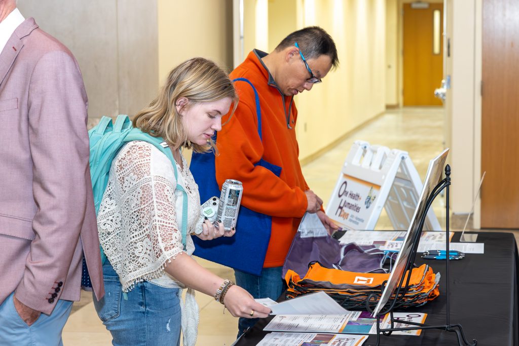 A blond young woman in a white lace-sleeved top and a man in a red pullover, glasses, and carrying a blue bag, stop to look at swag and flyers laid out on a table