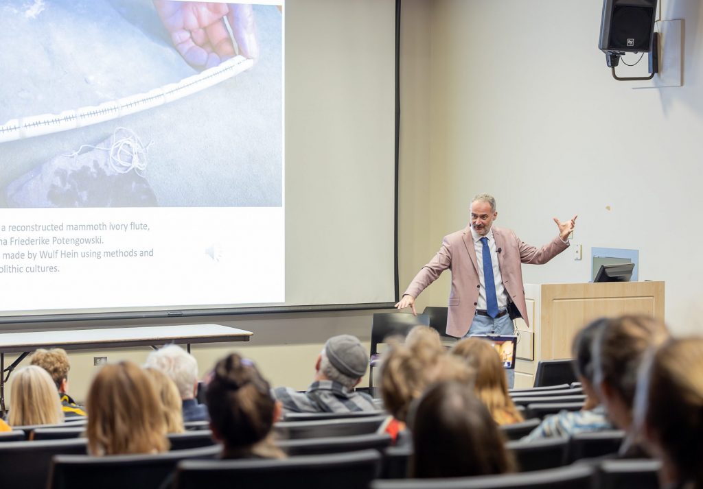 A white man in a pink blazer and blue tie gestures in front of a screen showing a slide of a person's hands holding an ancient bone recorder. In the foreground, from behind, are the heads of people in the audience listening to him speak.