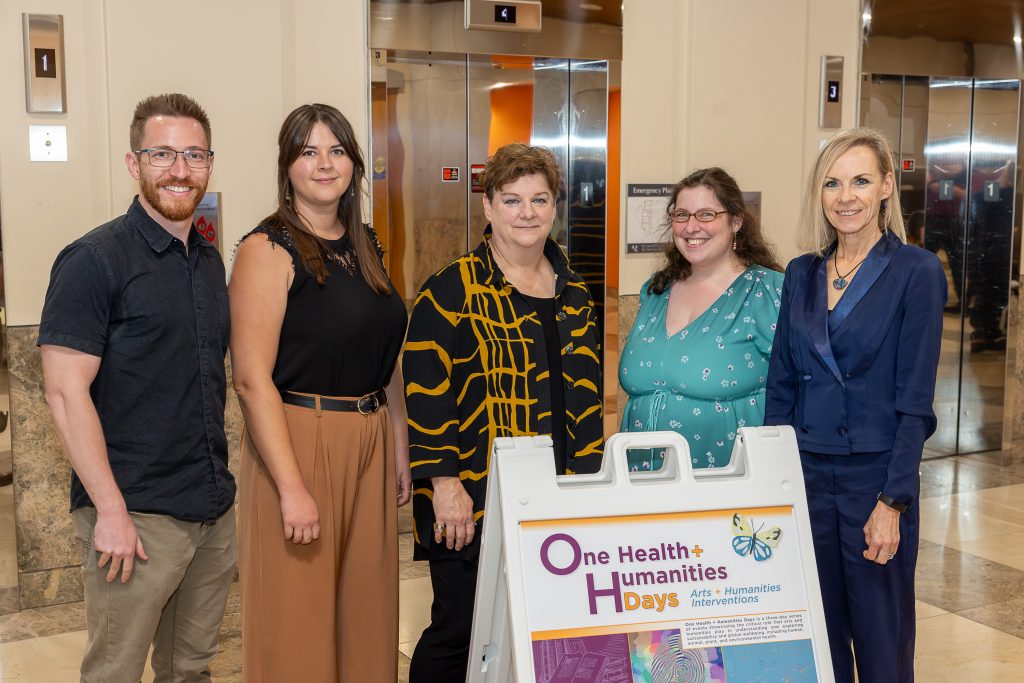 Photo of five people–a bearded white man and four women (two older, two younger) stand behind an A-frame sandwich board that reads "One Health + Humanities Days"