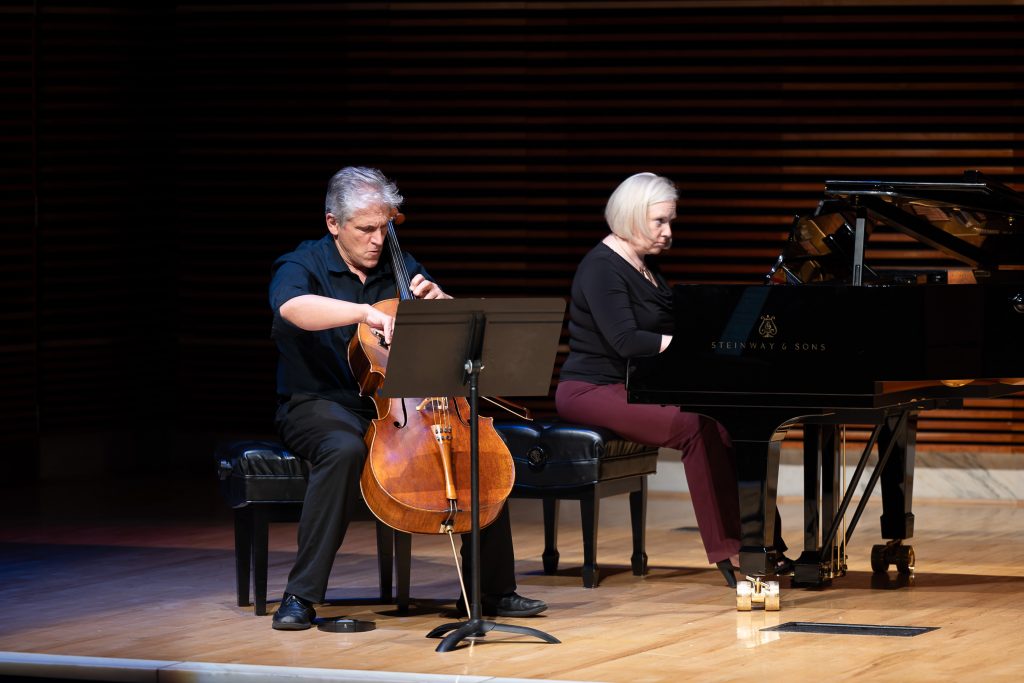 A photo of two people on a stage in a music hall. On the left is a man with short gray hair, seated on a bench playing the cello. On the right is a woman with shoulder-length gray hair playing a grand piano.