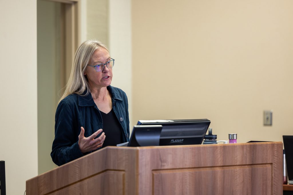 A white woman with long gray-blond hair and wearing a navy collared button-up over a black shirt stands behind a lectern giving a lecture