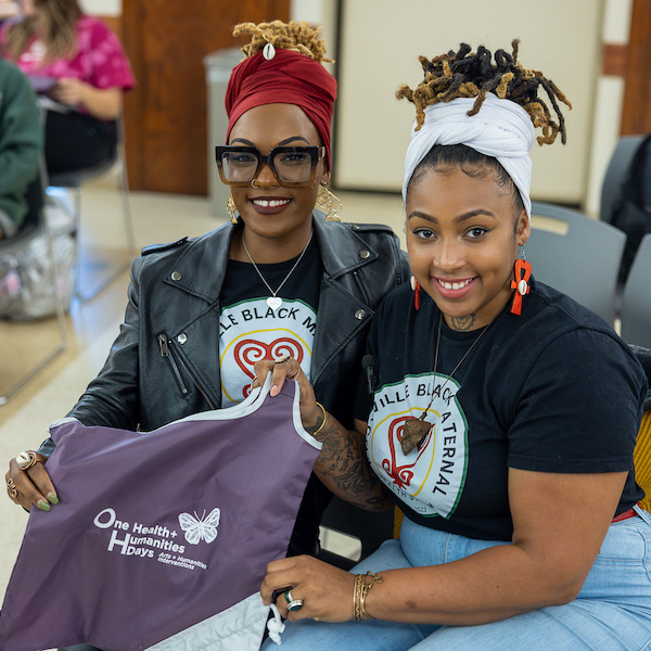 Two Black women sit smiling at the camera. One holds up a purple "One Health + Humanities Days" bag.