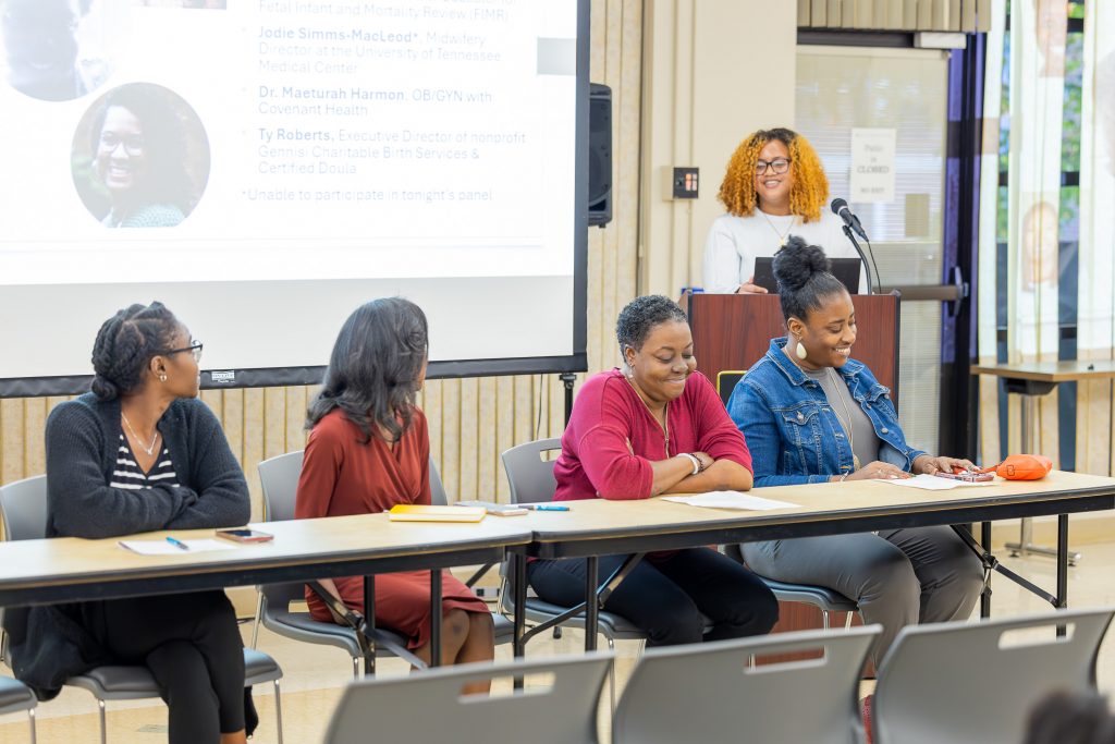 Four Black women sit smiling at a long conference table. Two of them are looking up at a fifth Black woman with orange hair who is standing behind a lectern.