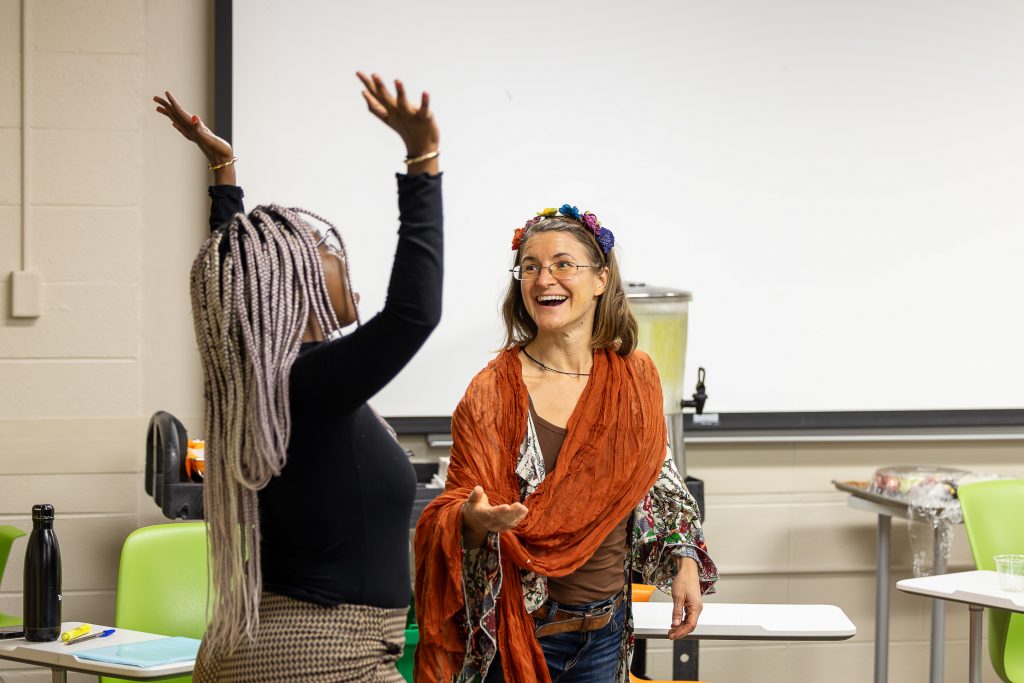 A Black woman with dreadlocks is on the left, shown from behind lifting her arms skyward in celebration. To her right is a white woman with brown hair wearing a flower wreath and red shawl, smiling happily as she reaches out to the first woman.