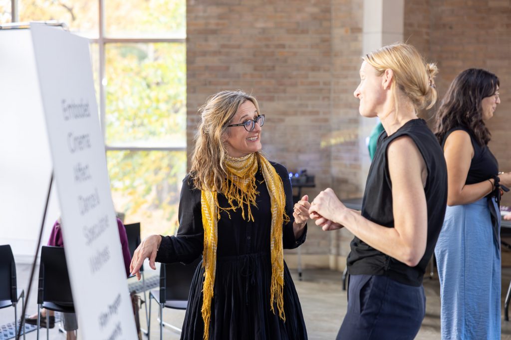 Two white women in dark clothing (one wearing a yellow scarf and glasses) talk animatedly with each other.