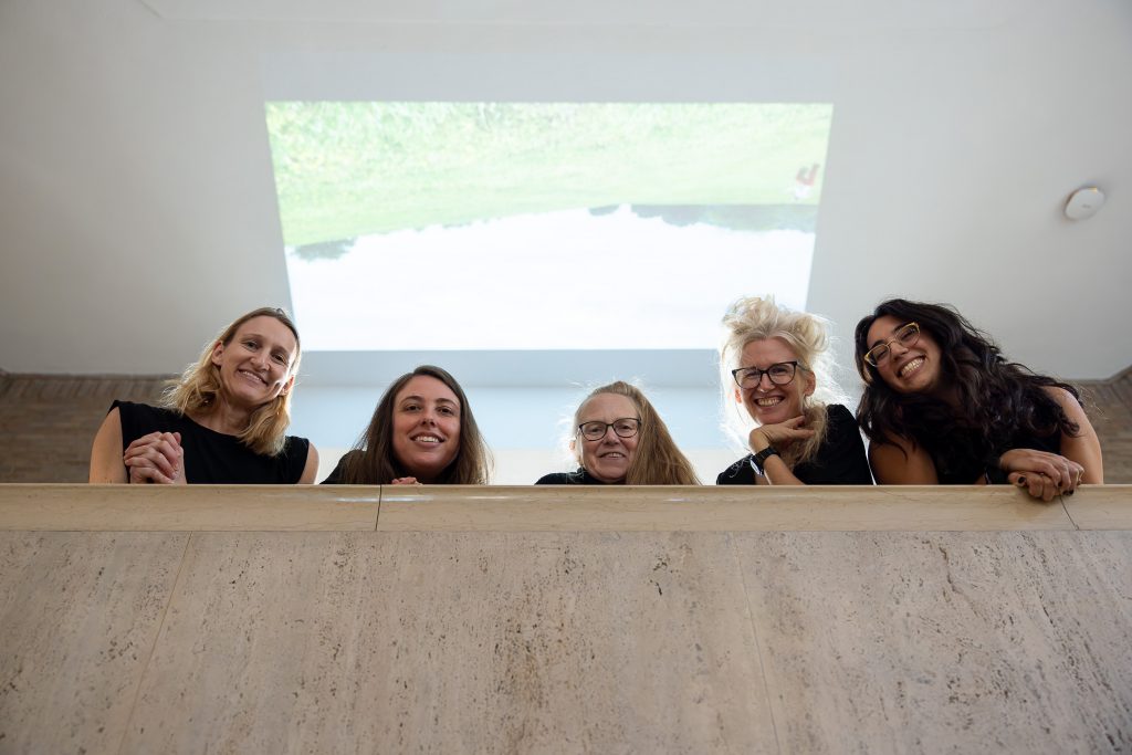 Five smiling white women look down from over a stone museum balcony. Above and behind them, projected on the ceiling, is an upside-down image of a green field with a wide sky over it.