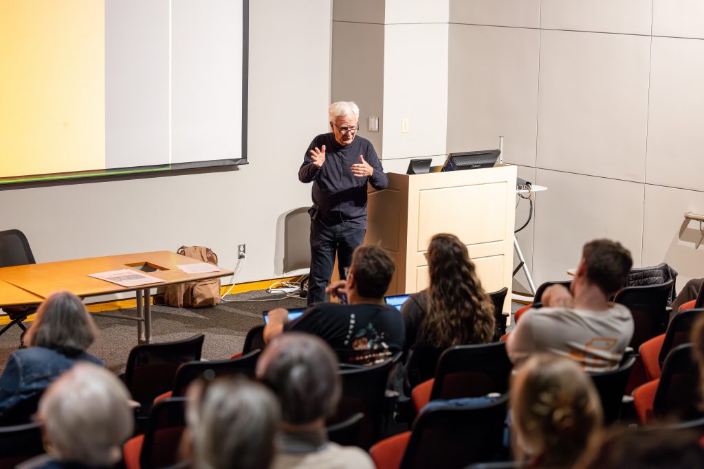 An older white man with dark-rimmed glasses stands in front of an audience in an auditorium giving a lecture