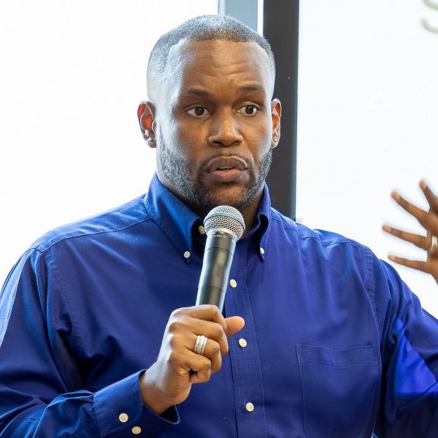 A Black man with short-cropped hair and beard gestures with one hand while holding a microphone in the other. He is wearing a blue button-up shirt.