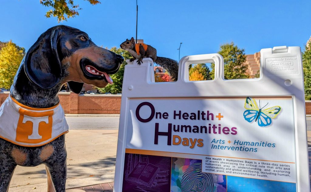 A statue of Smokey the bluetick hound dog mascot of UT is next to an A-frame sandwich board advertising One Health + Humanities Days. On top of the A-frame is a real live squirrel wearing an orange vest. The sky behind is blue.