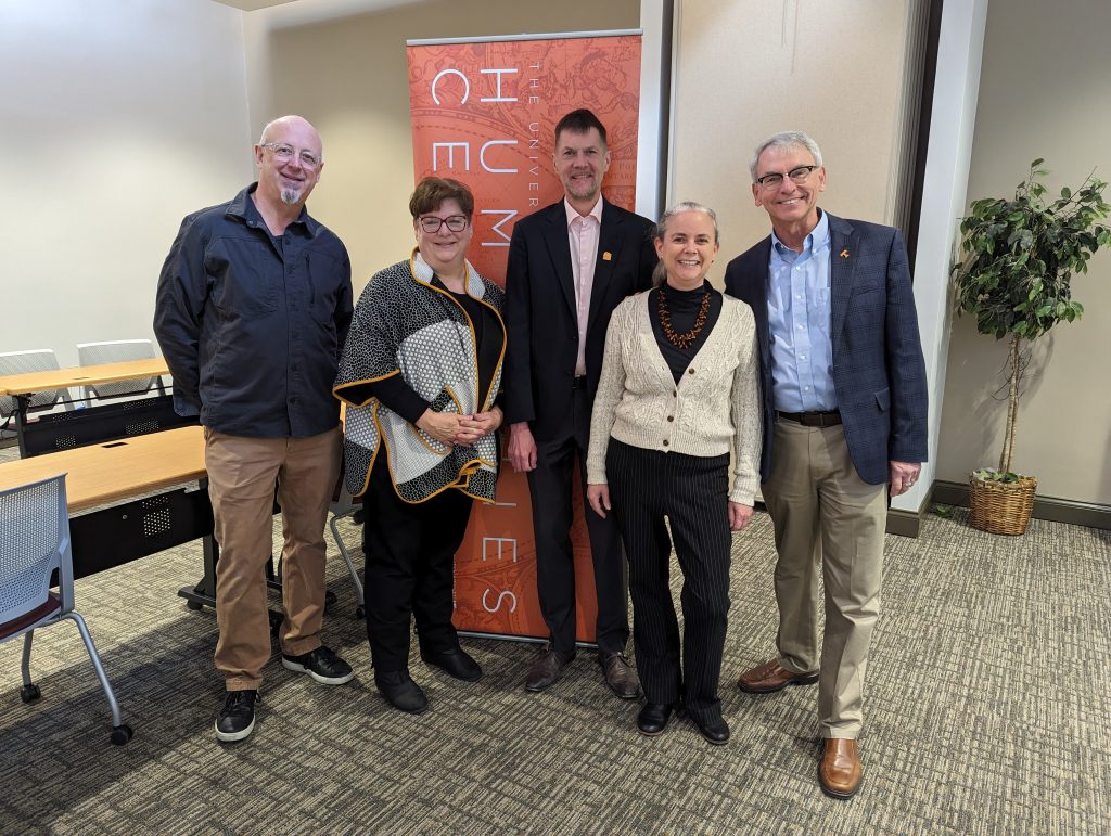 Five middle-aged white people in business clothes stand in front of a Humanities Center banner and smile at the camera