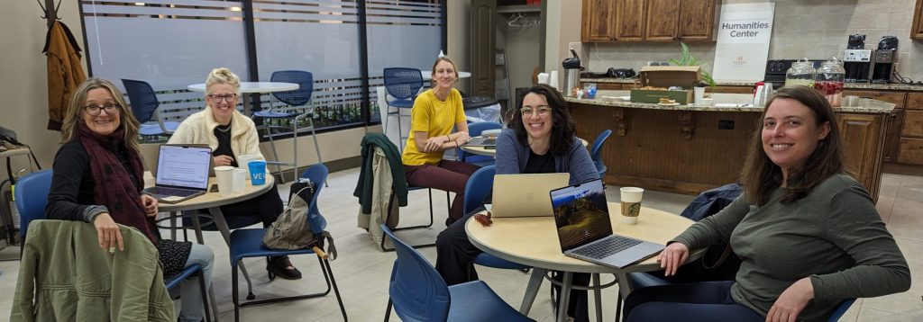 A photo shows a group of five smiling women sitting around several round tables in an office kitchen