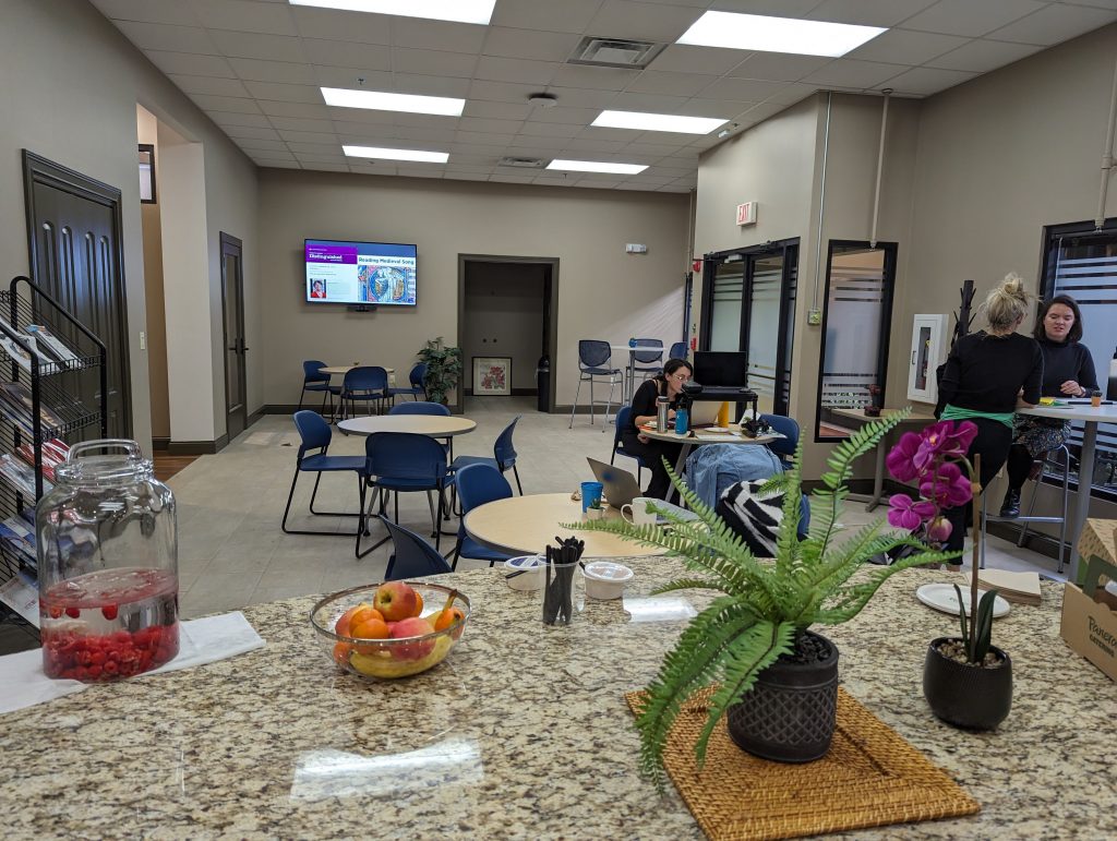 A large office kitchen contains several cafe tables with blue chairs, a few of which are occupied by some women working on computers and chatting. In the foreground is a granite countertop with some plants and food on it.