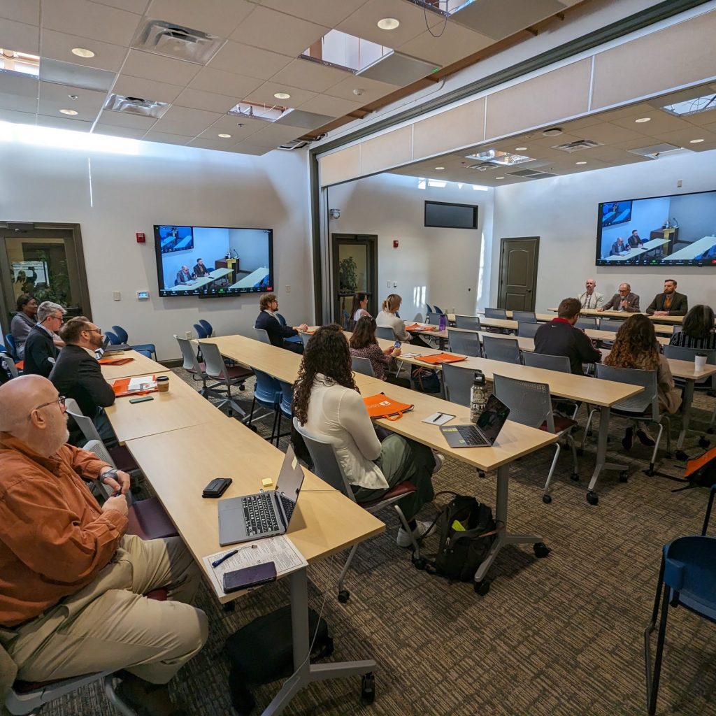 A room contains several rows of tables with people sitting at them, listening to three men seated at a front table. On the walls are two wall-mounted computer displays.