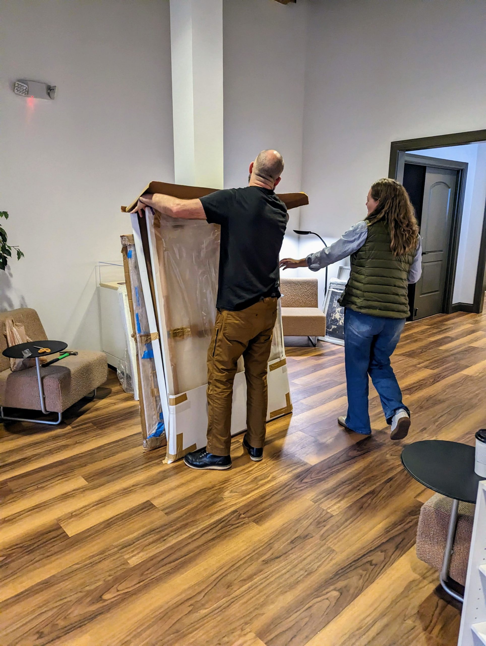 A balding man and a brown-haired woman unwrap large canvases covered in cardboard and plastic in preparation for hanging them.