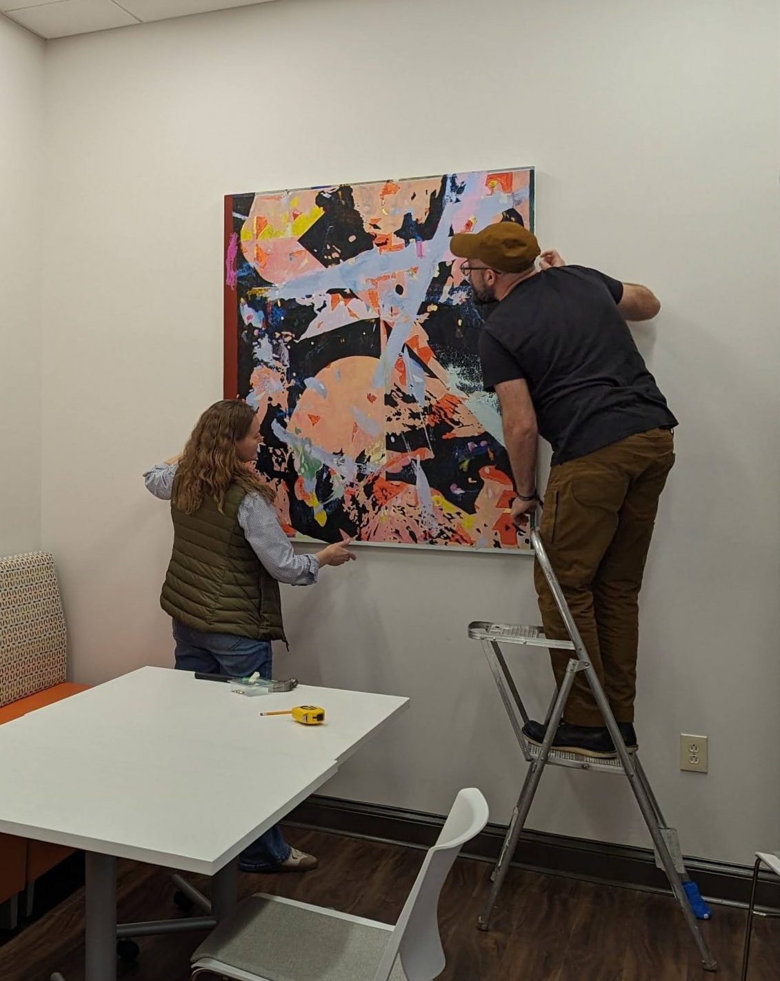 A woman in a vest with long brown hair helps a man in canvas pants and a matching brown canvas ball cap hang a large abstract painting on a wall. He is standing on a short stepladder. 