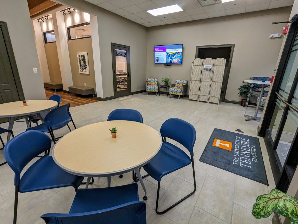 A large room leads off to a hallway on one side, and a set of double-doors with a UT floor rug in front of them on the other. In the foreground are a couple of round cafe tables with blue chairs around them.