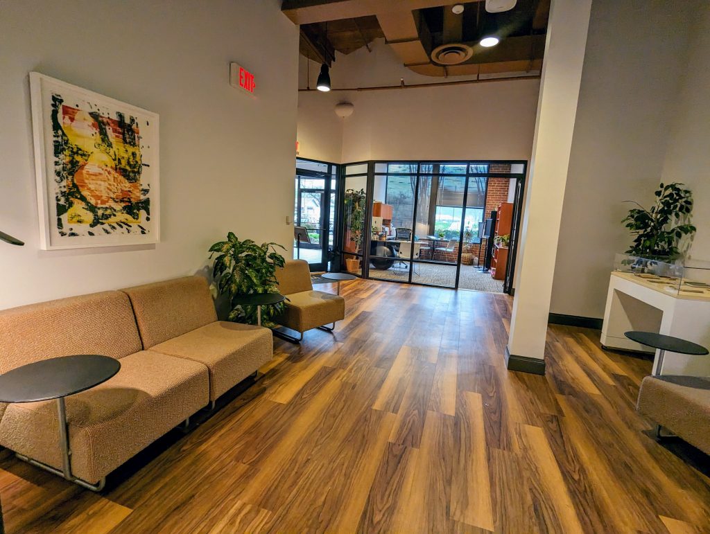 A high-ceilinged office building has brown wood-laminate floors leading to a glassed-in office and set of exterior doors. A pillar in the middle of the hall stretches up to to a ceiling with exposed duct work in brown. On either side of the hall are chairs and a small sofa, with a painting on the wall and some plants flanking them.