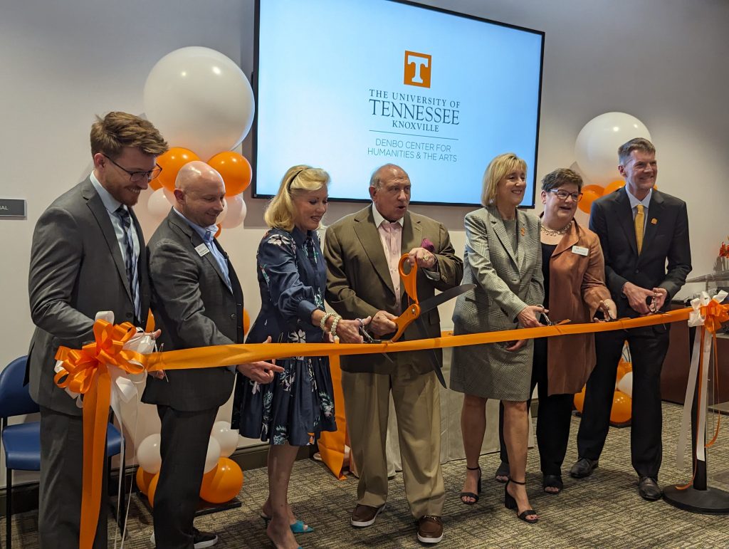 A group of seven well-dressed people stand in a row behind an orange ribbon strung between two posts. The older balding man in the center holds an oversized pair of orange-handled scissors in preparation for cutting the ribbon. Behind them on a call-mounted computer screen is a display of the Denbo Center's logo.