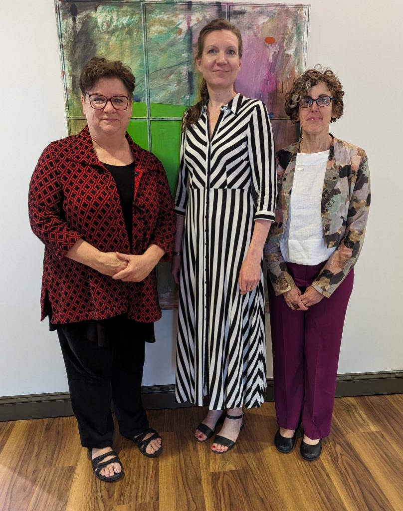 Three women stand in front of a modern painting. The woman on the left is wearing black slacks and a dark red dress jacket. The woman in the center-the tallest of them-wears a black-and-white striped long dress. The woman on the right is in eggplant-colored slacks, a white shirt, and a floral dress jacket.