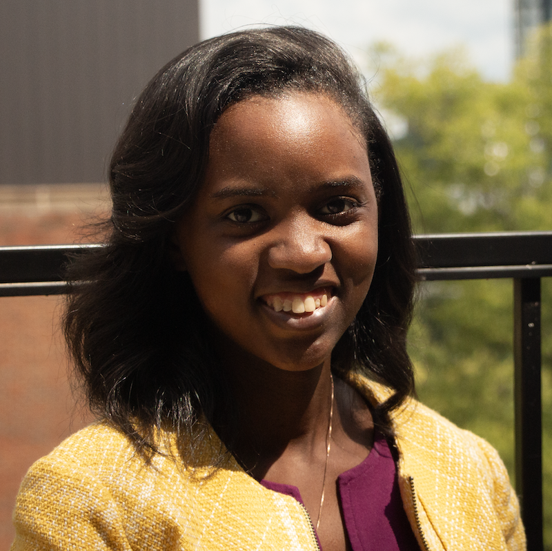 Headshot of a Black woman with shoulder-length straightened hair standing outside and wearing a yellow dress jacket over a maroon dress