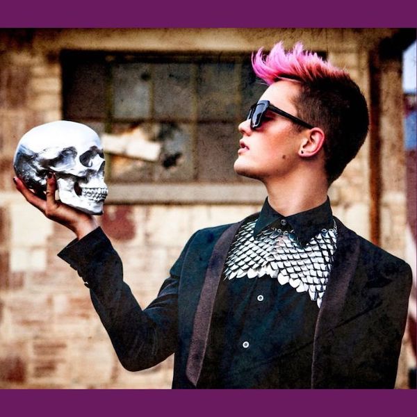 A white woman dressed in black with short, spiky pink hair and sunglasses holds and contemplates a human skull