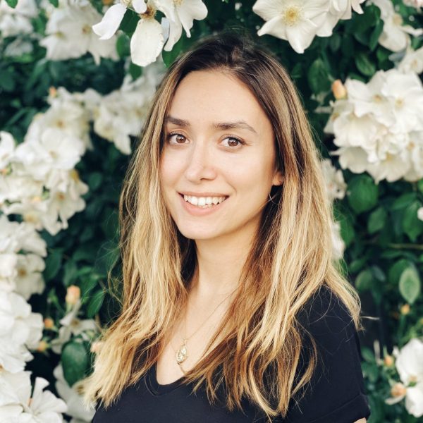 A photo of a young white woman with blond hair and dark highlights smiling at the camera. She is standing in front of a wall of plants with clusters of white flowers.